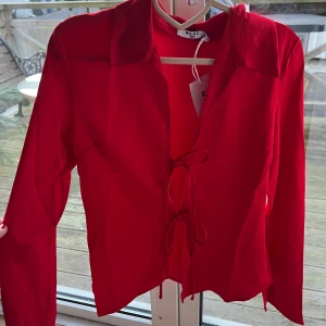 Röd satinblus från NA-KD - Snygg röd blus i satin från NA-KD med knytning framtill. Perfekt för att ge en färgklick till din outfit. Blusen har långa ärmar och en elegant design som passar till många tillfällen.