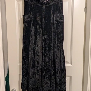 Svart sammet klänning - Elegant svart klänning i mjuk sammet med spetsdetaljer vid fållen. Klänningen har en dragkedja framtill och en vacker, flödande design som ger en lyxig känsla.