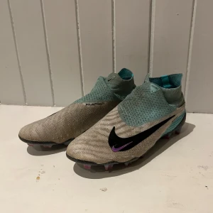 Nike phantom gx elite fg  - Snygga Nike fotbollsskor i en kombination av beige och turkos med en svart Nike-logga. Skorna har en strumpliknande passform och är designade för optimal prestanda på planen. Säljer på grund av att skorna inte passade min fot. 