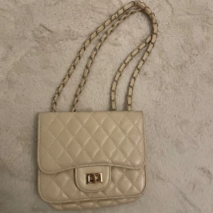 Beige quiltad axelväska - Snygg beige axelväska med quiltat mönster och kedjeaxelrem. Väskan har en praktisk dragkedja inuti och en elegant vridlåsdetalj på framsidan. Perfekt för att ge en stilren touch till din outfit.
