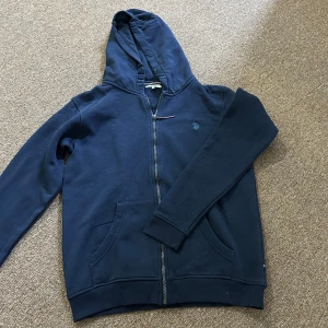 Mörkblå hoodie från U.S. Polo Assn. - Säljer en mörkblå hoodie från U.S. Polo Assn. med dragkedja och huva. Tröjan har långa ärmar och ribbade muddar vid ärmslut och nederkant. Perfekt för en avslappnad stil.