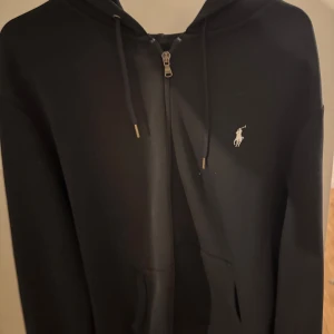 Svart hoodie från Ralph Lauren - Säljer en jättefin svart hoodie från Ralph Lauren med dragkedja och broderad logga på bröstet. Tröjan har långa ärmar och en praktisk huva med snören. Perfekt för en avslappnad look. Skicket är i 8/10 trots att tröjan är mer än 5 år gammal den har varit vältagen om och använt sparsamt ända anledningen det inte är en 10/10 är på grund av att den svarta färgen är inte lika mörk.