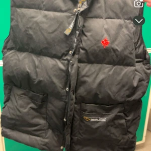Svart dunväst från Canada Goose - Säljer en svart dunväst från Canada Goose med en röd lönnlövdetalj på bröstet. Västen har en dragkedja och knappar framtill samt två stora fickor. Perfekt för kyliga dagar.