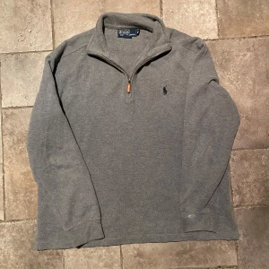 Ralph Lauren Quarter Zip Medium - Säljer en stilren grå Half/Quarer Zip från Polo Ralph Lauren i 100% bomull. Tröjan har en dragkedja vid halsen och det klassiska logotypbroderiet på bröstet. Perfekt för en avslappnad look. I riktigt gott skick utöver en mindre defekt på ena ärmen.