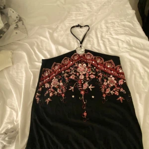 Svart topp med broderade detaljer - Elegant svart topp med vackra röda och vita broderade blommor framtill. Toppen har en halterneck-design med en dekorativ ringdetalj vid halsen. Perfekt för en speciell kväll!