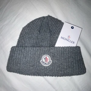 Moncler mössa  - Snygg grå ribbad mössa från Moncler med broderad logga framtill. Perfekt för kyliga dagar och ger en stilren look. Mössan är mjuk och bekväm att bära.