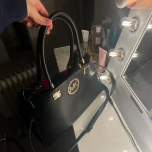 Svart handväska. ”MK” - Svart handväska med guldiga detaljer och logotyp framtill.  Den är inte äkta, köpt utomlands. Dragkedjan är trasig inuti väskan, annars är den i bra skick.  Säljer den då den inte kommer till användning.  Kontakta mig vid frågor eller för fler bilder.