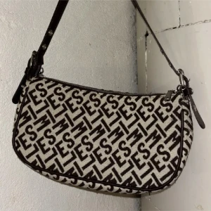 Mönstrad axelväska med kedjedetalj - Snygg axelväska med ett unikt mönster i svart och beige. Väskan har en kedjedetalj och justerbar axelrem i mörkt skinn. Perfekt för att ge din outfit en trendig touch.