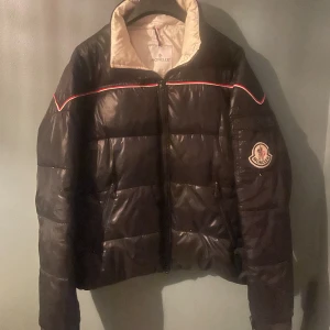 Vintage svart pufferjacka från Moncler - Säljer en svart pufferjacka från Moncler med dragkedja och broderad logga på ärmen. Jackan har en quiltad design och är tillverkad i syntetmaterial. Den har en snygg detalj med röd och vit rand längs axlarna. Inga defekter. Priset är inte fast. Kan byta. Kan mötas i Gbg, annars står köparen för frakten!