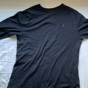 Svart långärmad tröja från Ralph Lauren - Säljer en stilren svart långärmad tröja från Ralph Lauren med det klassiska röda logotypen på bröstet. Perfekt för en avslappnad och snygg look. Tröjan är tillverkad i mjukt bomullsmaterial.