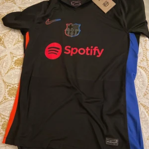 Svart FC Barcelona fotbollströja från Nike - Snygg svart FC Barcelona tröja med korta ärmar och färgglada detaljer. Tröjan har klubbens emblem och Spotify-logga på framsidan samt UNHCR-logga på baksidan. Tillverkad i ett lätt och bekvämt material, perfekt för fotbollsfans. Den ör helt ny köpte den men den var stor på mig. Om man köper båda real Madrid t shirt och den så får man båda för 800 och med samma leverans så man inte betalar för bådas leverans. Priset kan diskuteras