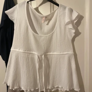Vit topp från Nelly - Säljer en söt vit topp från Nelly i storlek M. Den har korta ärmar och en volangdetalj vid ärmslut och nederkant. Toppen är ribbad och har en knytning i midjan för en fin passform. Perfekt för sommardagar eller en avslappnad vårlook.