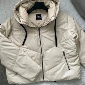 Beige pufferjacka från Zara - Säljer en snygg beige pufferjacka från Zara. Jackan är perfekt för kyliga dagar och har en mysig huva med svarta snören. Den är i mycket bra skick och passar perfekt för höst och vinter.