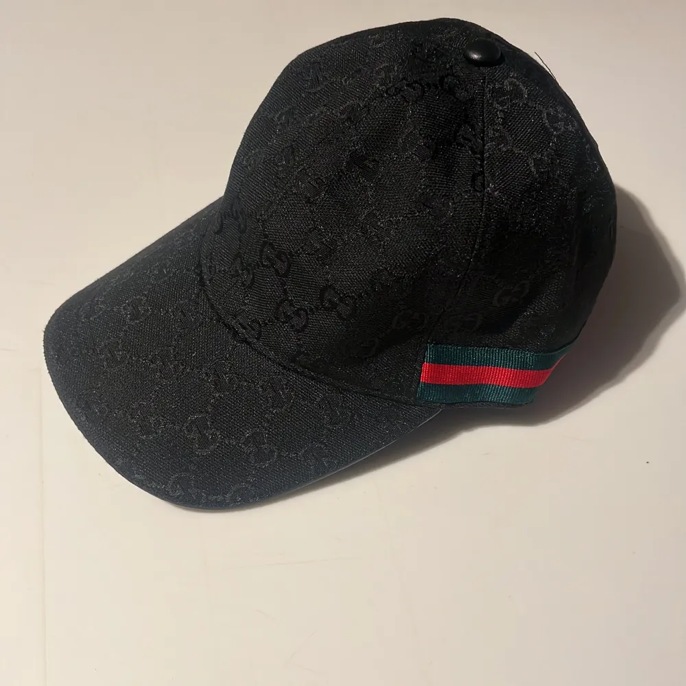 Snygg svart keps med diskret mönster och en randig detalj i grönt och rött på sidan. Kepsen har en justerbar rem baktill för perfekt passform.🧢✔️⚫️⚫️. Accessoarer.