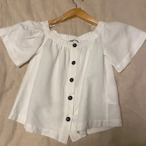 Vit blus från H&M - Säljer en söt vit blus från H&M i storlek XS. Den har en offshoulder-design med korta ärmar och knappar framtill. Perfekt för sommardagar eller en avslappnad vårlook. Blusen är i mycket bra skick och redo för nya äventyr!