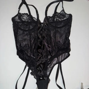 Svart spetskorsett med snörning - Snygg svart korsett i spets med justerbara axelband och snörning framtill. Perfekt för en festlig kväll eller en speciell tillställning. Den har en elegant och sexig design med genomskinliga detaljer.