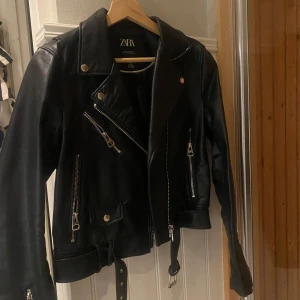 Svart skinnjacka från Zara - Snygg svart skinnjacka från Zara med dragkedjor och bälte. Perfekt för en cool look med sina detaljer och klassiska bikerstil. Jackan har långa ärmar och en stilren design. passar mig som oftast ha S