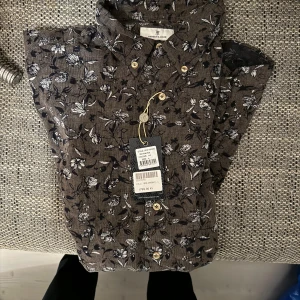 Helt ny Brun blommig skjorta från H&H - Snygg brun skjorta från H&H med ett stilrent blommönster i vitt och svart. Skjortan har knappar framtill och är perfekt för en trendig look.