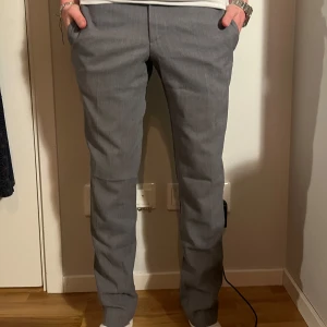 Grå kostymbyxor från J.Lindeberg - Snygga grå kostymbyxor från J.Lindeberg i mycket bra skick. De har en klassisk straight passform och är perfekta för både kontoret och finare tillfällen. Byxorna har knappar och dragkedja framtill samt fickor på sidorna. Perfekt för höst och vår!
