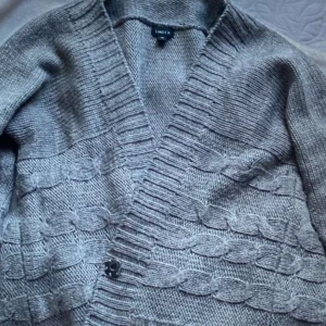 Grå stickad kofta från Indiska - Mysig grå stickad kofta från Indiska med ett snyggt flätmönster. Den har en djup v-ringning och knappar framtill. Perfekt för kyliga dagar när du vill vara både varm och stilren. Passar bra till både vardag och lite finare tillfällen.