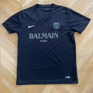 Svart PSG x Balmain fotbollströja - Snygg svart fotbollströja från Nike i samarbete med PSG och Balmain. Tröjan har korta ärmar och en stilren design med Balmain Paris-logga på bröstet. 