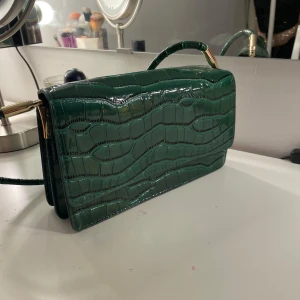 Grön axelväska med krokodilmönster - Snygg grön axelväska med krokodilmönster och gulddetaljer. Perfekt för att ge en touch av elegans till din outfit. Väskan har en stilren design och en praktisk axelrem.