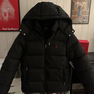 Svart dunjacka från Polo Ralph Lauren - Snygg svart dunjacka från Polo Ralph Lauren med huva och dragkedja. Jackan har ett broderat rött märke på bröstet och praktiska fickor med dragkedjor. Perfekt för kyliga dagar.