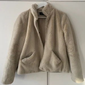 Beige teddyjacka från Only - Mysig beige teddyjacka från Only med knappar. Jackan har långa ärmar och två fickor framtill. Perfekt för kyliga dagar när du vill hålla dig varm och stilren. 