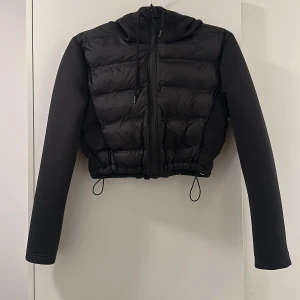 Svart croppad pufferjacka med huva - Snygg svart croppad pufferjacka med huva och dragkedja. Jackan har långa ärmar och justerbar dragsko i nederkant för en perfekt passform. Perfekt för kyliga dagar när du vill hålla stilen.
