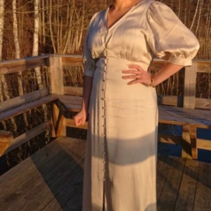 Beige långklänning med puffiga armar  - Säljer en elegant beige långklänning med puffiga trekvartsärmar och knappar framtill. Klänningen har en v-ringad design och är perfekt för vår och sommar. Materialet känns mjukt och bekvämt, vilket gör den idealisk för både fest och vardag. Endast använt en gång , prislapp kvar . Orginalpris: 799