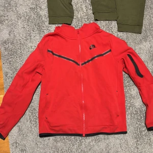 Röd  Nike tech fleece - Säljer en röd hoodie från Nike i bra skick. Den har en dragkedja framtill och en huva. Perfekt för höst och vår. Några små fläckar finns, men inget som stör. Passar både till träning och vardag. Pris diskuteras vid snabb affär