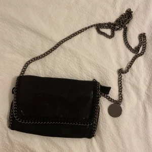 Svart axelväska med kedjedetaljer - Snygg svart axelväska med coola kedjedetaljer runt kanterna och en lång kedjerem. Perfekt för en utekväll eller fest! Väskan har en stilren design och är i mycket bra skick. Den är liten och smidig, perfekt för att bära med sig det viktigaste.