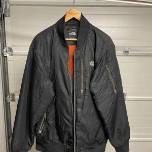 Svart bomberjacka från The North Face - Säljer en snygg svart bomberjacka från The North Face i bra skick. Den har en klassisk design med dragkedja och ribbade muddar. Insidan är orange, vilket ger en cool kontrast. Perfekt för höst och vår! Säljes i befintligt skick. Storlek: XL