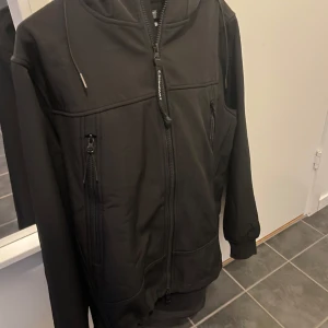 Cp company jacka - Säljer en svart cp company jacka i mycket bra skick. Den har en dragkedja framtill och praktiska fickor med dragkedjor. Perfekt för höst och vår, med en bekväm passform och långärmade ärmar. Huvan ger extra skydd mot väder och vind. Perfekt för vardagsbruk!