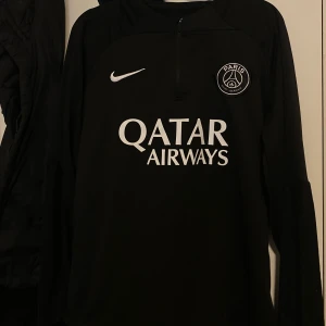 Svart långärmad fotbollströja från Nike - Snygg svart långärmad fotbollströja från Nike med Paris Saint-Germain-logga och Qatar Airways-tryck på framsidan. Perfekt för träning eller som supportertröja. Enkel och stilren design med dragkedja vid halsen. Den säljer jag pga att den aldrig kommer till användning längre men skicket är 10/10