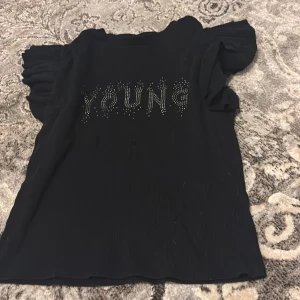 Svart topp med volangdetaljer - Snygg svart topp med texten 'YOUNG' i glittriga stenar på framsidan. Toppen har volangdetaljer vid ärmarna som ger en trendig look. Perfekt för att piffa upp vardagsstilen!