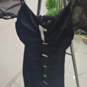 Svart klänning med knappar pris kan diskuteras - Elegant svart klänning med knappar framtill. Den har en stilren design och är perfekt för en kväll ute. Klänningen har korta ärmar med transparenta detaljer som ger en extra touch.