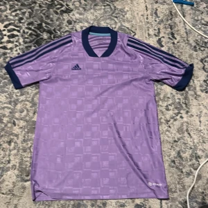 Lila fotbollströja från Adidas - Snygg lila fotbollströja från Adidas med mörkblå detaljer och AEROREADY-teknologi. Tröjan har korta ärmar och ett diskret mönster som ger en stilren look. Perfekt för både träning och match! Pris kam diskuteras.