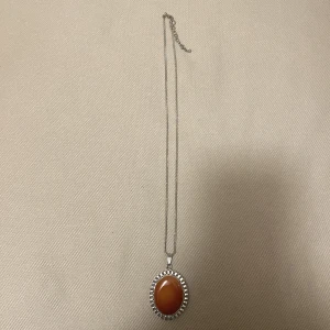 Silverhalsband med orange sten - Säljer ett elegant silverhalsband med en stor oval orange sten i mitten. Stenen är omgiven av en detaljerad silverram som ger en vintagekänsla. Kedjan är tunn och stilren, vilket gör att hänget verkligen står ut. Perfekt för att ge en färgklick till din outfit!