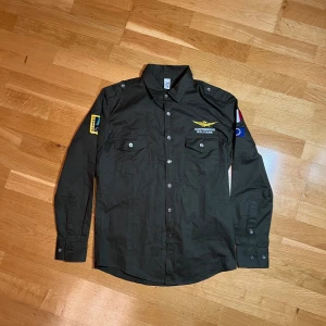 Militärgrön skjorta med broderade märken - Säljer en italienska flygvapnets militärgröna skjorta med broderade märken på ärmarna. Skjortan har långa ärmar och knappar framtill. Perfekt för en avslappnad stil eller temafest. Märkena inkluderar italienska flaggan och flygvapnets logga Frecce Tricolori. Skjortan är i bra skick och redo för nya äventyr! Sitter mer som en smal. 