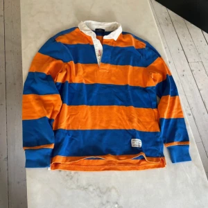 Randig pikétröja från GANT - Säljer en cool randig pikétröja från GANT i blått och orange. Tröjan har en klassisk krage och knappar framtill. Perfekt för en avslappnad stil och passar bra till både jeans och chinos. Den är i bra skick och redo för nya äventyr!