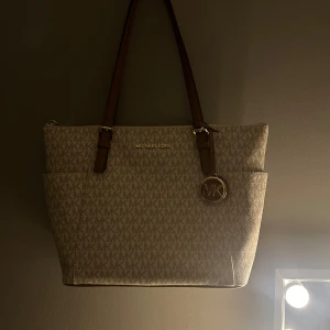 Beige axelväska från Michael Kors - Snygg beige axelväska från Michael Kors med det klassiska MK-mönstret. Väskan har bruna axelremmar och en dekorativ MK-berlock. Perfekt för både vardag och fest! Aldrig använts!