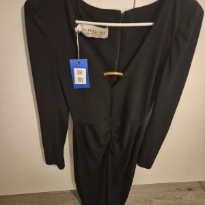 Svart långklänning från By Malina - Elegant svart långklänning från By Malina i nytt skick. Klänningen har en v-ringad design och långa ärmar, perfekt för höst och vinter. Materialet känns lyxigt och passar utmärkt för festliga tillfällen. Den har en stilren och tidlös look som gör den lätt att matcha med olika accessoarer.