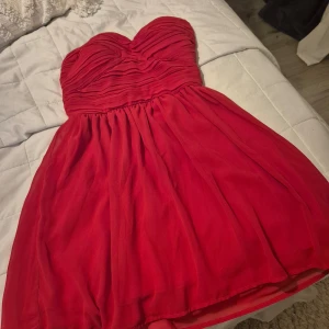 Röd axelbandslös klänning - Säljer en superfin röd axelbandslös klänning med rynkad överdel. Perfekt för fest eller nyår! Klänningen har en härlig passform och är i mycket bra skick. Den är kort och har en vacker, flytande kjol.