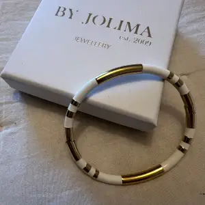 Snyggt armband från By Jolima i vitt och 14K guldplätterat stål. Det har en stilren design med växlande sektioner av vit och guld. Perfekt för att ge en elegant touch till vilken outfit som helst. Kommer i originalförpackning.