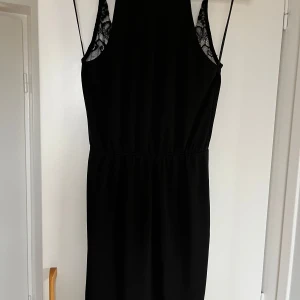 Svart klänning med spetsdetaljer - Elegant svart klänning med vackra spetsdetaljer vid axlarna och en djup v-ringning. Perfekt för festliga tillfällen. Klänningen har en smickrande passform och är ärmlös, vilket gör den idealisk för både sommar och nyår.