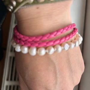 Rosa flätat armband med pärlor - Snyggt armband med en mix av rosa flätat band och vita Sötvattenpärlor . Armbandet har en silverfärgad kedja och ett litet runt hänge. Perfekt för att ge en färgklick till din outfit!