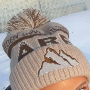 Beige Åre mössa - Superfin beige Åre mössa med bergsmotiv och en stor tofs på toppen. Perfekt för kalla vinterdagar. Den har en ribbad kant och text i brunt och vitt. Passar perfekt nu inför vintern!