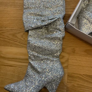Glittriga silverstövletter NLY SHOES - Säljer ett par fantastiska glittriga stövlar i silver. De har en spetsig tå och en elegant klack som ger en extra touch av glamour. Perfekta för festliga tillfällen när du vill stå ut och glittra lite extra. Skorna är använda en gång och kommer i originalförpackning. Se bild för att se vad som hänt med ena klacken