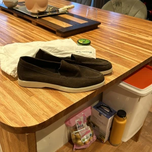 Bruna mockaloafers - Säljer ett par snygga bruna loafers i mocka. De har en stilren design med vit sula som ger en fräsch kontrast. Perfekta för både vardag och fest! Skorna är i mycket bra skick och kommer med en praktisk tygpåse för förvaring. Dom har aldrig använts utom hus